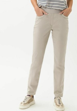 Beige Hose mit glatter Textur, ausgestattet mit seitlichen Taschen und einer engen Passform. Kombiniert mit hellen Sneakers für einen lässigen Look.
