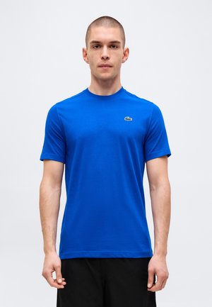 Hombre con cabello corto que lleva una camiseta de manga corta azul real lisa con un pequeño logo de cocodrilo verde en el pecho izquierdo.