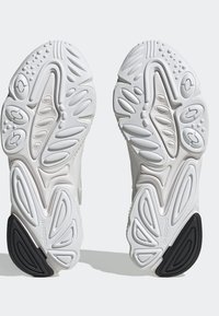 Vita atletiska skosulor med texturerade gummimönster, som kombinerar släta och räfflade områden. Svart detalj på hälen.