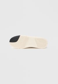 Semelle de chaussure présentant une base en caoutchouc beige avec une texture antidérapante, un accent noir au niveau du talon, et un motif de semelle pour une meilleure traction.