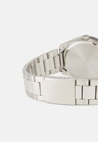Montre-bracelet en acier inoxydable argenté avec un bracelet à maillons métalliques, un boîtier rond lisse et un fermoir simple. Logo de la marque visible sur la boucle.