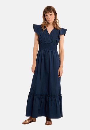 Donna che indossa un abito maxi blu navy con maniche arricciate, vita arricciata e orlo a balze, abbinato a sandali bassi marroni.