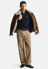 Bruine shearling jas, marineblauwe trui, gestreept overhemd, beige broek en donkere schoenen. Opvallende details: zachte voering, contrasterende kraag en riem.