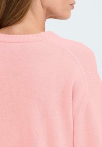 Ljust rosa stickad tröja med ribbad rundhals och raglanärmar. Texturen ser mjuk och något lös ut.