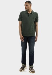 Polo shirt vert olive, à manches courtes, col à trois boutons. Associé à un jean bleu foncé et des chaussures de sport marron avec des accents jaunes.