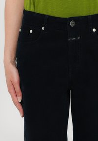 Gros plan d'une personne portant un pantalon en velours côtelé noir et un haut vert, une main reposant à côté de la poche du pantalon.