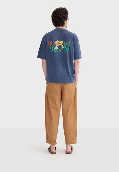 Homme aux cheveux bouclés portant un t-shirt bleu à motif floral, un pantalon ample beige et des sandales, debout de dos, les mains dans les poches.