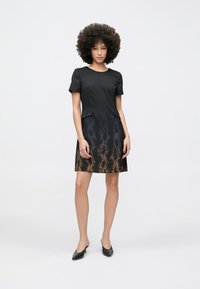 Femeie cu păr creț purtând o rochie neagră cu mâneci scurte și imprimeu de șarpe multicolor în partea de jos, stând în pantofi negri cu toc.