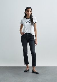 T-shirt branca de mangas curtas com gola preta e gráfico "MOTHER", combinada com jeans escuros justos e sapatilhas pretas, sobre um fundo neutro.