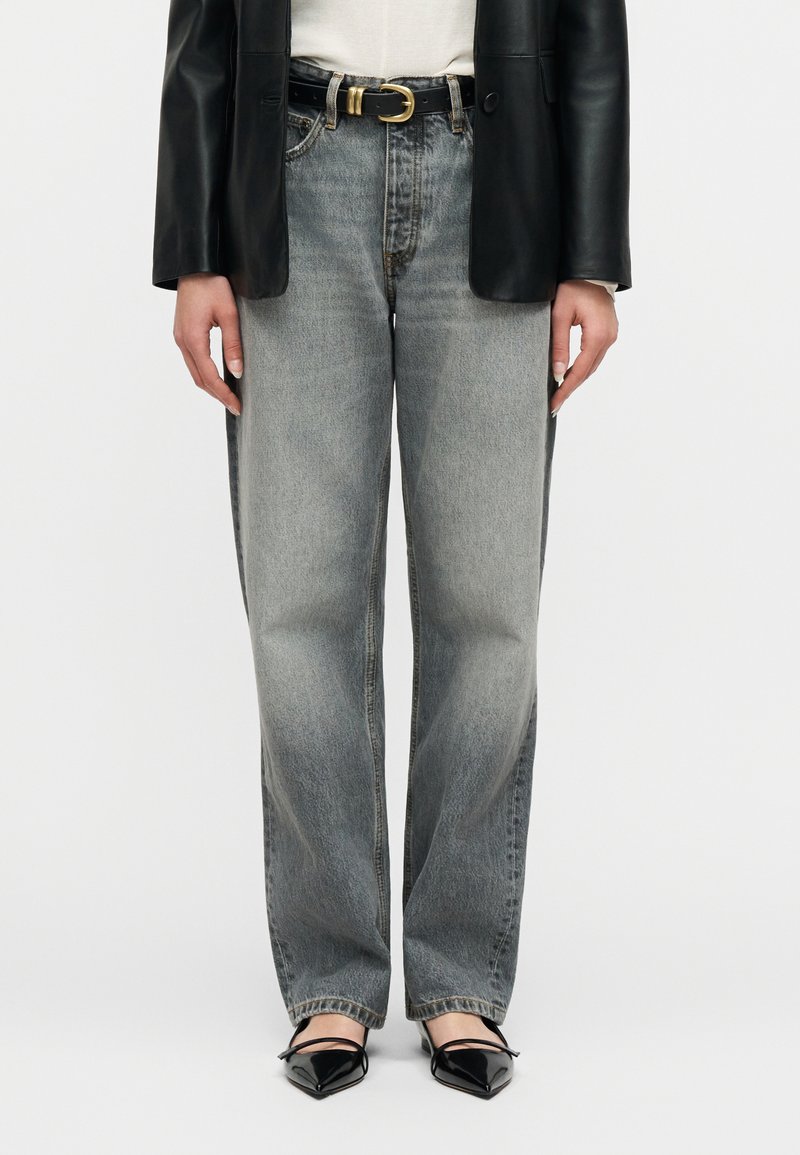 Άτομο που φοράει ξεβαμμένο τζιν κα νονικού μήκους, μαύρη δερμάτινη ζώνη με χρυσή αγκράφα, μαύρο δερμάτινο σακάκι, λευκό τοπ και μαύρα μυτερά παπούτσια.