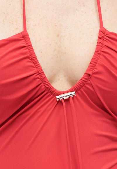 Κοντινή λήψη από κόκκινο πάνω μέρος μαγιό με ρυθμιζόμενα λεπτά λουράκια και μικρή ασημένια πόρπη στη V-σχήματος λαιμόκοψη.