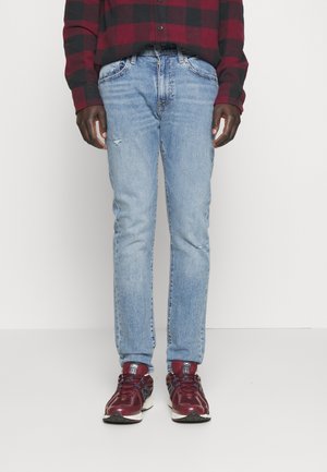 Personne portant un jean bleu délavé, des baskets bordeaux et une chemise à carreaux rouge et noir, debout devant un fond blanc uni.