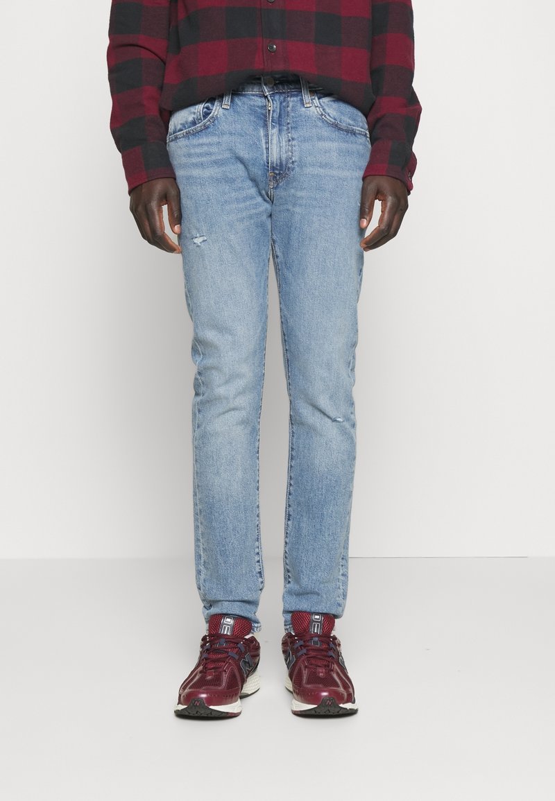 Personne portant un jean bleu délavé, des baskets bordeaux et une chemise à carreaux rouge et noir, debout devant un fond blanc uni.
