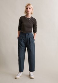 Brązowa bluzka z długim rękawem, ciemnoturkusowe spodnie z wysokim stanem i plisami oraz białe tenisówki tworzące swobodną sylwetkę na neutralnym tle.