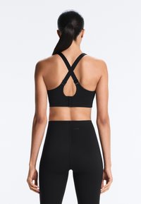 Ženska z dolgimi temnimi lasmi, spetimi v čop, oblečena v črno športno nedrček z začinjenimi naramnicami in visokim pasom črne legice, stoji obrnjena stran.