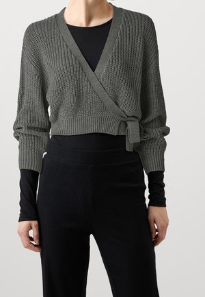 Femme portant un pull court en maille gris à enfiler et noué à la taille, par-dessus un haut noir à manches longues et un pantalon côtelé noir.