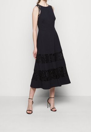 Femme portant une robe midi noire sans manches avec des panneaux en dentelle et des sandales noires à talons hauts avec bride à la cheville, debout devant un fond clair uni.
