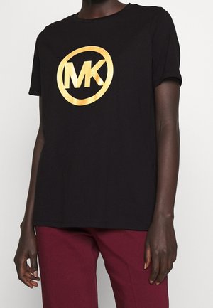 Persoon in een zwart t-shirt met een groot gouden "MK" logo en burgundy broek, gefotografeerd van de nek tot halverwege de dij.