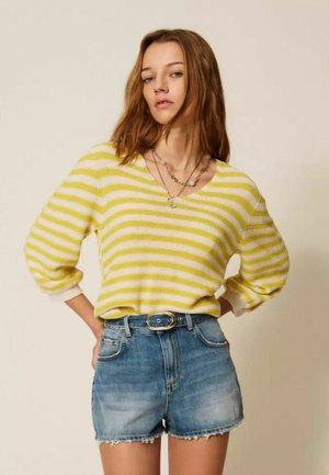 Giovane donna con capelli castani fino alle spalle che indossa un maglione a righe giallo e bianco, shorts in denim blu e collane a strati, in piedi con le mani dietro.