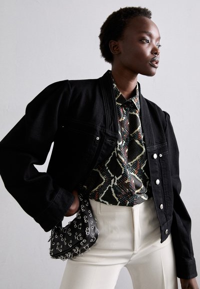 Giacca di jeans nera con chiusura a bottoni, camicia decorata sotto, pantaloni bianchi su misura e una piccola borsa nera con un design argentato.