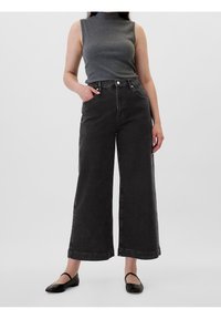 Jean en denim noir à jambes larges avec une taille haute, associé à un haut gris sans manches. La texture est lisse, avec des poches standards et des accessoires métalliques.