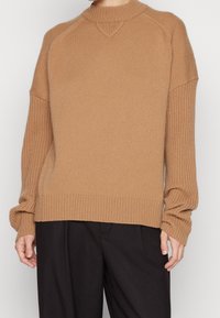 Pull marron avec une texture côtelée, col montant et épaules tombantes. Présente une coupe ample et des poignets étroits côtelés.