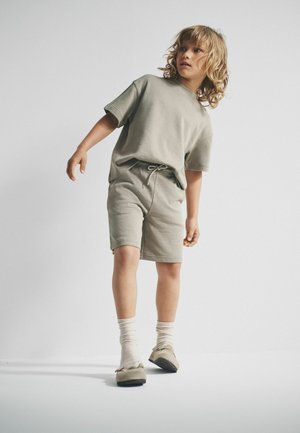 Child with curly blonde hair wearing beige oversized T-shirt, matching shorts, white socks, and beige shoes standing against plain background.