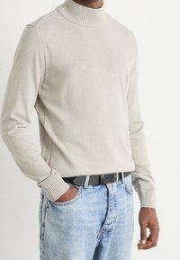 Pull à col roulé beige clair avec poignets et col côtelés, associé à un jean bleu délavé et une ceinture noire. Pas de motifs ni logos visibles.