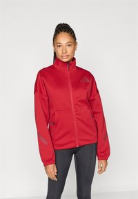 Červená bunda na zips vyrobená z hladkého, matného materiálu. Má vysoký golier, klokanie vrecká a reflexné prvky na rukávoch.