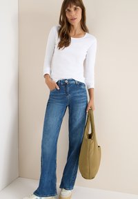 Hvid langærmet skjorte, blå flagrende jeans og en grøn taske. Sneakers med beige accenter. Enkel design, afslappet udseende og løs pasform.