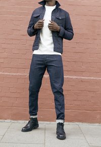 Denimjacka i marinblått med knäppt front, vit t-shirt med rund hals, mörka jeans och svarta läderkängor med snörning, stående mot en rosa vägg.