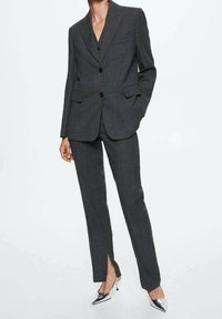 Femme portant un costume gris foncé sur mesure avec une veste boutonnée et un pantalon droit fendu à l'avant, assorti à des escarpins argentés à bout pointu.