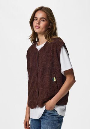 Jeune femme aux cheveux châtain clair portant une veste polaire marron par-dessus un t-shirt blanc et un jean bleu, debout avec une main dans la poche.