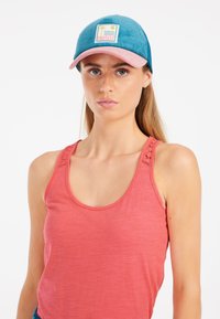 Blaues Cordhut mit einem rosa Schirm und einem Logo-Patch, das ein Sonnen- und Wassermotiv zeigt. Das Modell trägt ein korallfarbenes Tanktop mit detaillierten Trägern.