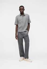 Γκρι καρό πουκάμισο με κοντά μανίκια και κουμπιά στο μπροστινό μέρος· σε συνδυασμό με άνετο γκρι cargo παντελόνι με πλαϊνές τσέπες. Λευκά αθλητικά παπούτσια ολοκληρώνουν την εμφάνιση.