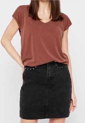 Kobieta w rdzawej, krótkiej bluzce z dekoltem w serek, wpuszczonej w wysokowatą czarną spódnicę z denimu z ćwiekowanymi kieszeniami.