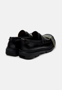 Scarpe slip-on in pelle nera con suola spessa, caratterizzate da una finitura liscia e un logo discreto sul lato. Dettagli di cucitura visibili.