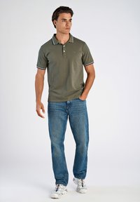 Olivgroen poloshirt met een drieknoops placket, witte strepen op de kraag en mouwen. Gecombineerd met een blauwe spijkerbroek en witte sneakers.