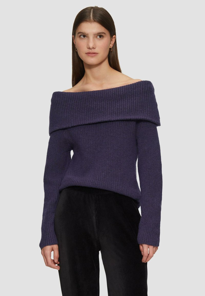 Schulterfreier lila gerippter Strickpullover mit langen Ärmeln, strukturiertem Stoff und entspanntem Schnitt, kombiniert mit schwarzen Cordhosen.