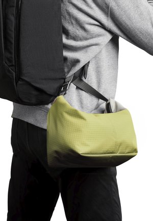 Grüne Tasche aus Verbundmaterial mit geometrischem Muster, getragen von einer Person in grauem Sweatshirt und mit einem schwarzen Rucksack.