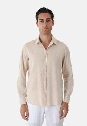 Uomo con capelli scuri che indossa una camicia beige a maniche lunghe con bottoni e pantaloni bianchi, in piedi davanti a uno sfondo chiaro e uniforme.