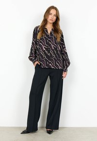 Svart blus med abstrakt mönster i kräm och lila, med V-ringning och lös passform. Matchad med svarta vida byxor och spetsiga skor.