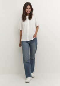 White, button-up blouse with short puff sleeves, paired with light blue straight-leg jeans and white sneakers on a neutral background.