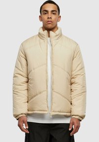 Beige polstret jakke med høy krage, med diagonale sømdetaljer og elastiske mansjetter. Glidelås foran, lagd over en hvit topp.