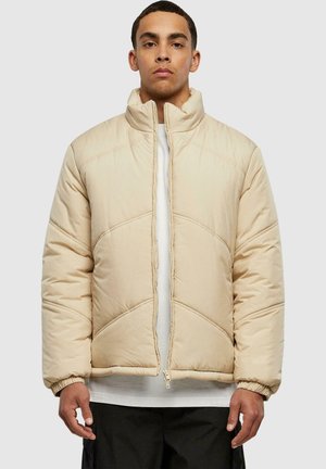 Beige padded jacket with a high collar, featuring diagonal stitching details and elastic cuffs. Zippered front closure, layered over a white top.