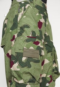 Cargo pants in green camo pattern with dark green and burgundy accents. Features multiple pockets and stitched details, made from cotton blend fabric.