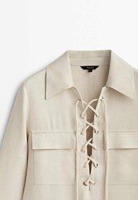 Beige Bluse mit Kragen, ausgestattet mit einer Schnürfront und zwei Brusttaschen. Hergestellt aus einem strukturierten Stoff mit einer weichen Oberfläche.