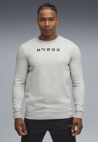 Sweatshirt en coton gris à manches longues, col rond, en tissu texturé, avec le logo noir "HYROX" sur la poitrine et des détails aux poignets.
