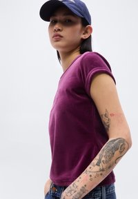 Νεαρό άτομο που φοράει πορφυρό t-shirt και μπλε καπέλο, με λεπτομερείς τατουάζ στο δεξί αντιβράχιό του και ουδέτερη έκφραση.