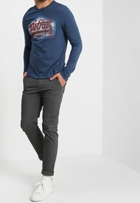 Námořnické tričko s dlouhým rukávem s potištěným logem; šedé slim-fit kalhoty; bílé tenisky. Materiál působí hladce a střih je volnější.